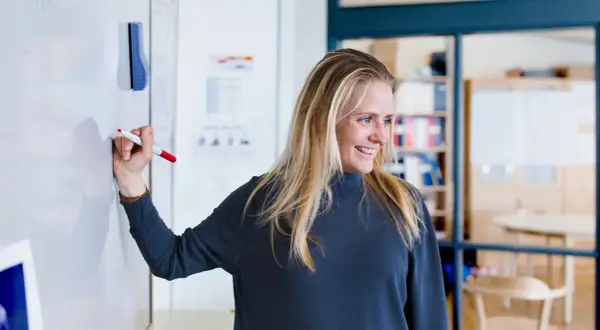 Glad kvinna med blondt hår skriver på en whiteboard. Hon kanske studerar socialpedagog utbildning distans.