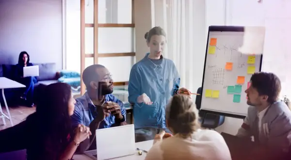 Mötesdeltagare diskuterar en presentation om hållbarhet, liknande en byggnadsantikvarie utbildning. En kvinna leder diskussionen vid en whiteboard.