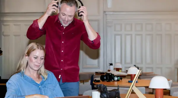 Man med hörlurar och kvinna vid laptop, en hundstatyett på bordet. De arbetar med sitt it konsult-projekt.
