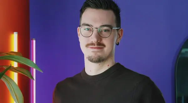 En ung man med glasögon och mustasch ler mot kameran. Bakgrunden är lila med en röd vägg och en växt. Han kanske är student på Chas Academy.