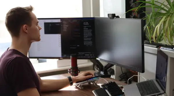 Man vid datorn, fokuserad på sin programmerings kurs. Flera skärmar visar kod och text. En mikrofon och en webbkamera är synliga.