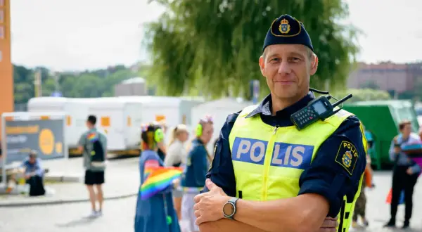 Polis i tjänst, kanske på väg att plugga till polis. Han bär gul reflexväst med texten "POLIS" och en keps med Sveriges riksvapen. I bakgrunden syns folk och en regnbågsflagga.
