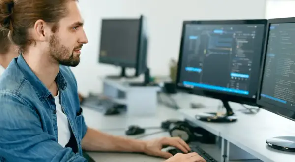 Manlig IT-tekniker med uppsatt hår och skägg arbetar vid två datorskärmar med kod.