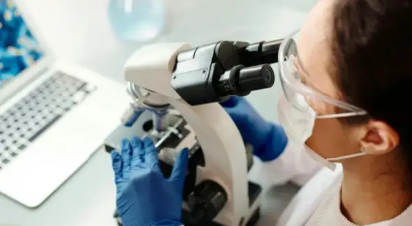 En forensiker arbetar med ett mikroskop i ett laboratorium. En laptop syns i bakgrunden.