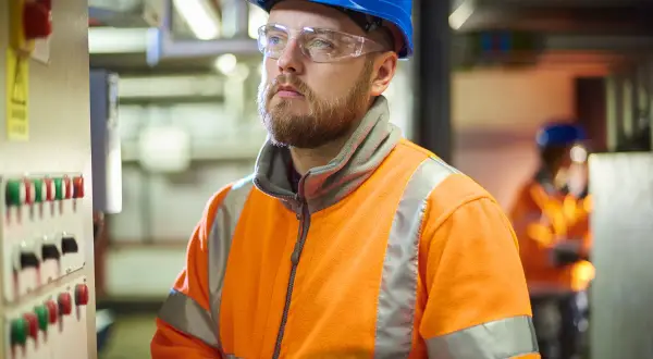 Man i blå hjälm och varselkläder vid kontrollpanel. En del av en produktionstekniker utbildning.