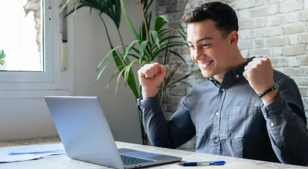 Ung man jublar framför sin laptop, kanske har han hittat roliga jobb eller lyckats med ett projekt.