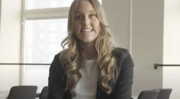 En ung kvinna med blont, lockigt hår ler och talar. Hon bär en svart kavaj över en vit topp. Bakom henne syns fönster och stolar, kanske i en lokal på Katedralskolan, Uppsala.