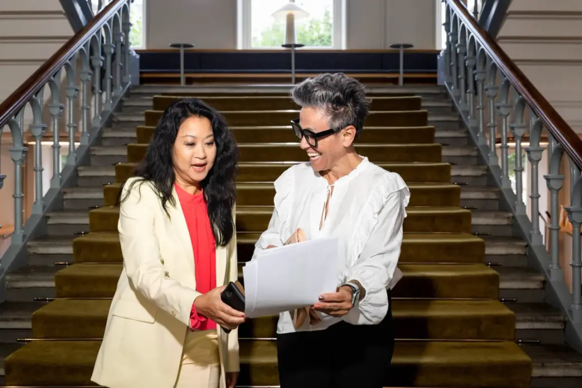 Två kvinnor diskuterar papper vid en trappa. Den ena kvinnan, klädd i ljus kavaj, ger en telefon till den andra, som ler och håller i dokumenten. Kanske en personlig assistent som hjälper till med organisationen.