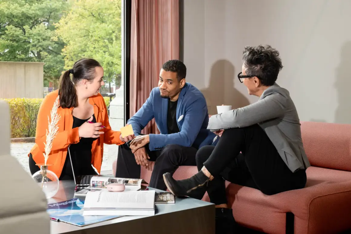 Tre personer diskuterar, en kvinna i orange ger en gul post-it till en man i blå kavaj. Kanske planerar de en virtuell assistent utbildning.