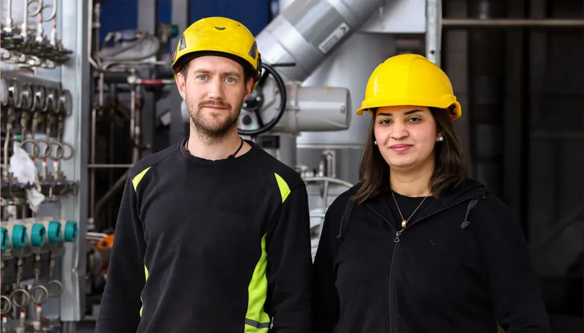 Två personer med gula hjälmar står framför industriell utrustning. De är studenter vid byggbranschens utbildningscenter.