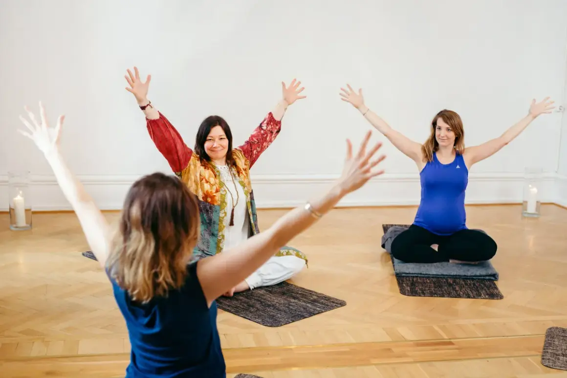 Tre kvinnor sitter på yogamattor med armarna uppåt. En av dem är gravid. En inspirerande scen från en yoga utbildning.