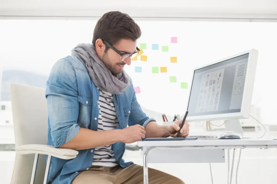 En grafisk designer arbetar vid sitt skrivbord med en ritplatta och dator. Färgade post-it-lappar sitter på väggen bakom.