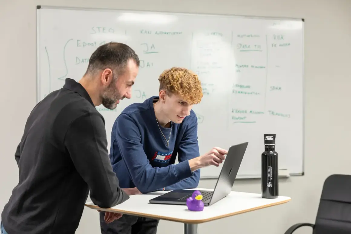 Två män diskuterar kod på en laptop. En .net utvecklare pekar på skärmen medan en kollega tittar på. En whiteboard med anteckningar syns i bakgrunden.