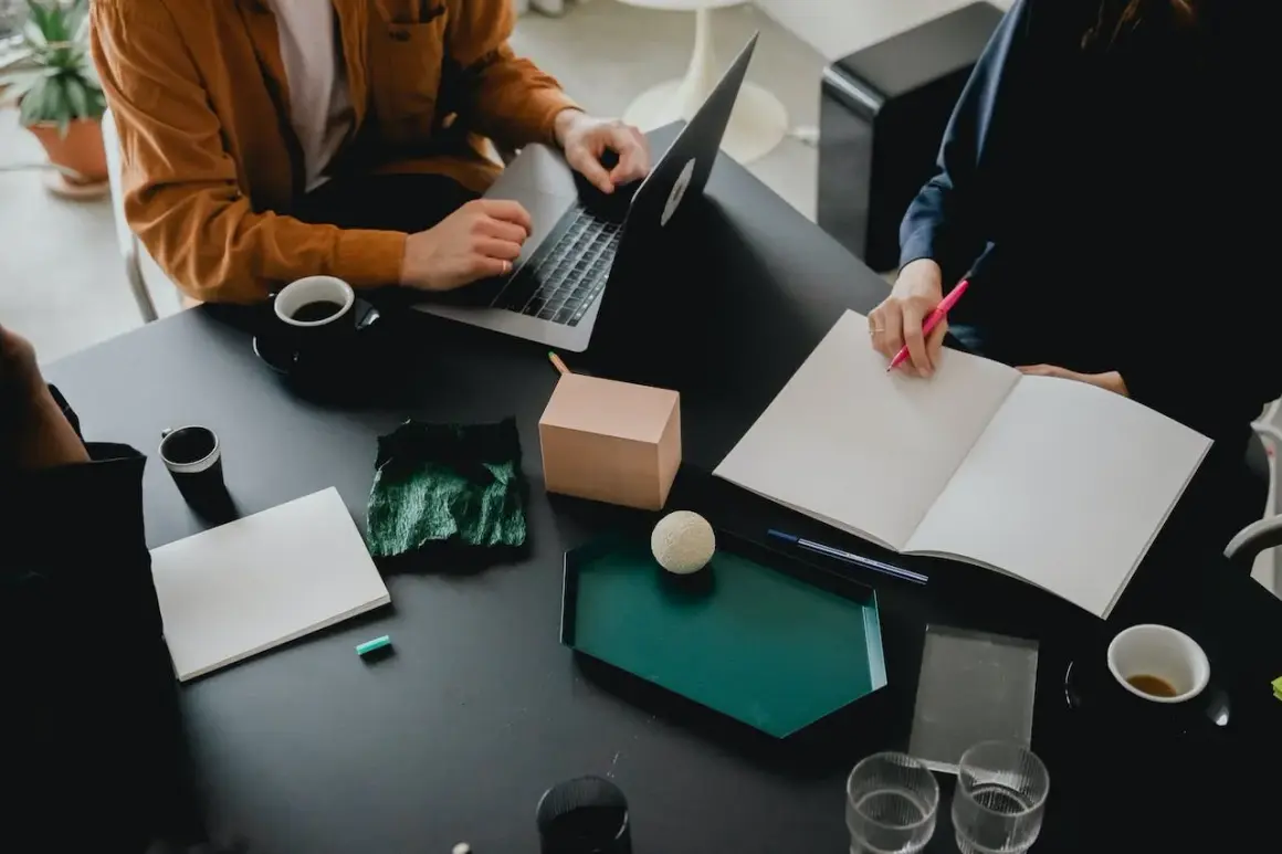 Två personer vid ett bord, en med laptop, den andra skriver i en bok. Kaffe och anteckningsmaterial. De är bi utvecklare som samarbetar.
