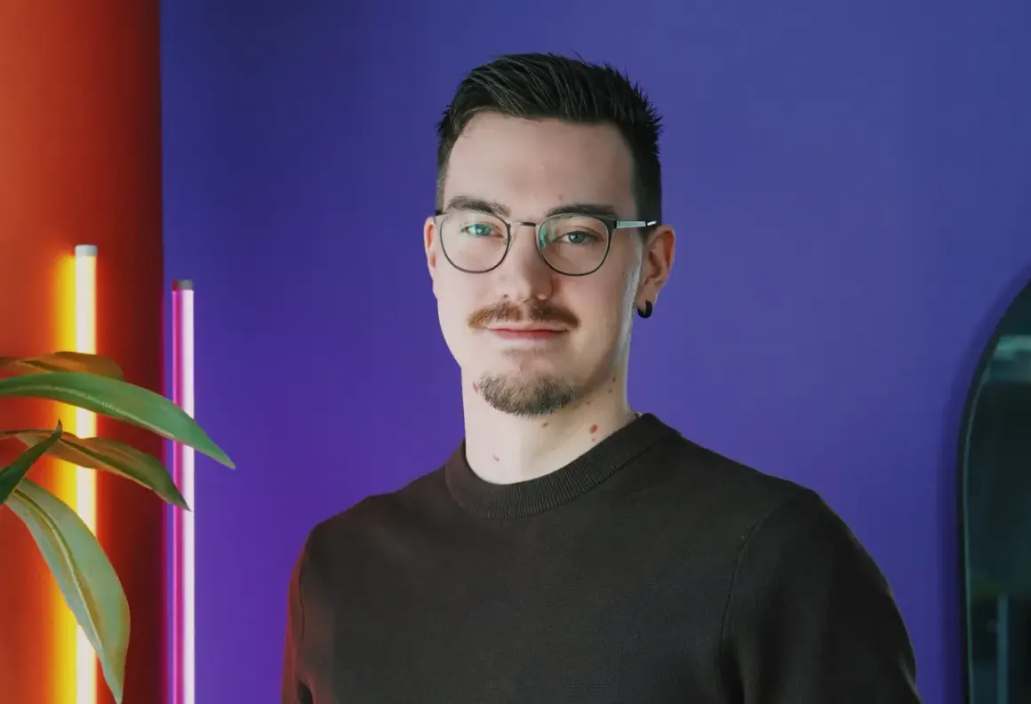En ung man med glasögon och mustasch ler mot kameran. Bakgrunden är lila med en röd vägg och en växt. Han kanske är student på Chas Academy.