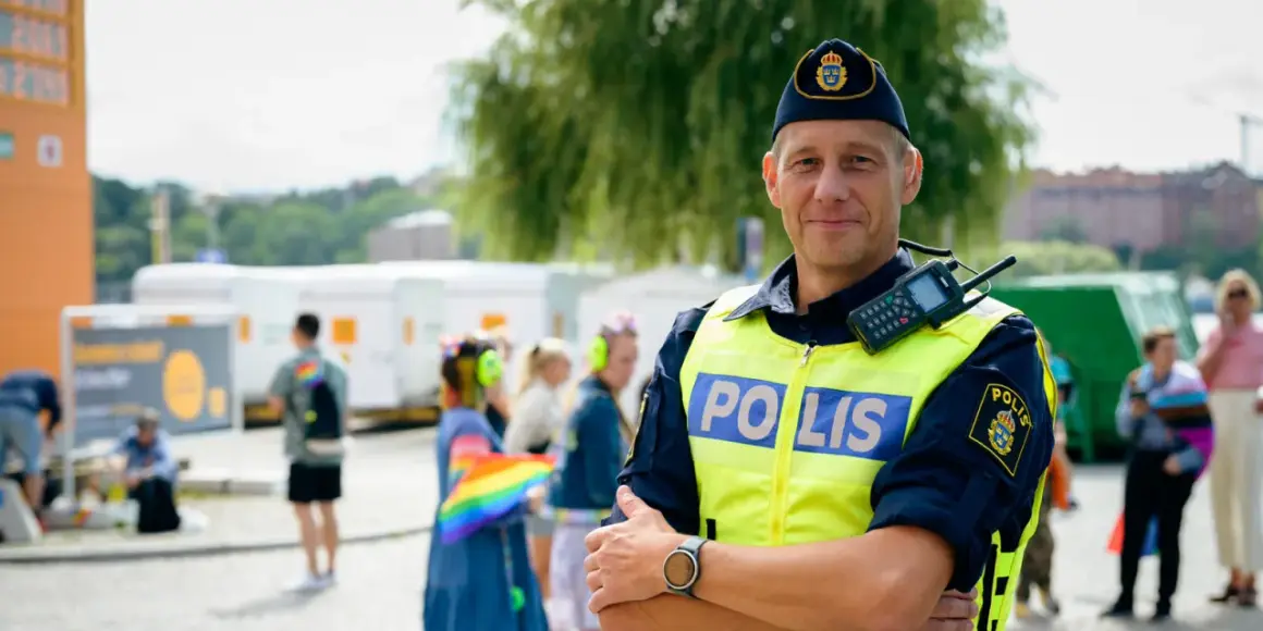 Polis i tjänst, kanske på väg att plugga till polis. Han bär gul reflexväst med texten "POLIS" och en keps med Sveriges riksvapen. I bakgrunden syns folk och en regnbågsflagga.
