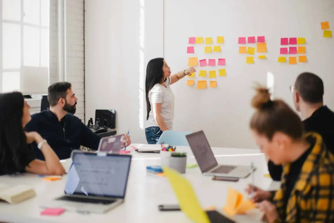 Teamwork i ett ljust kontor. En kvinna pekar på post-it-lappar på en vägg, medan andra diskuterar. Kanske en embedded utvecklare som presenterar idéer.