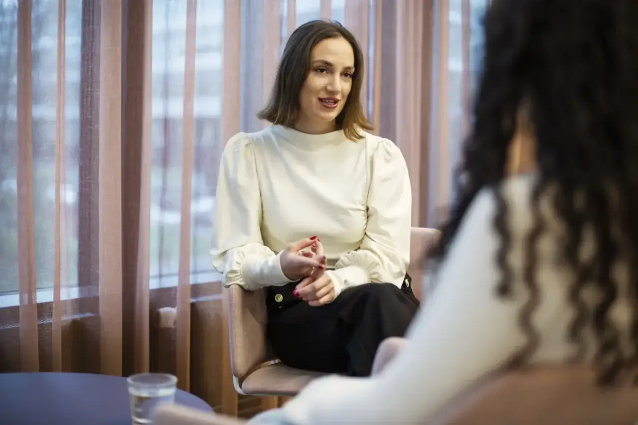 En kvinna med kort brunt hår och vit blus sitter i en stol och pratar. Hon verkar förklara psykolog utbildning krav.