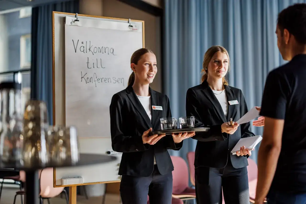 Tv&aring; studenter fr&aring;n hotell och turismprogrammet v&auml;lkomnar g&auml;ster till en konferens.