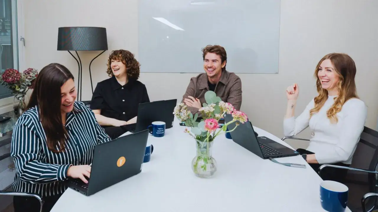 Ett team av glada .net utvecklare skrattar runt ett bord med laptops och blommor.