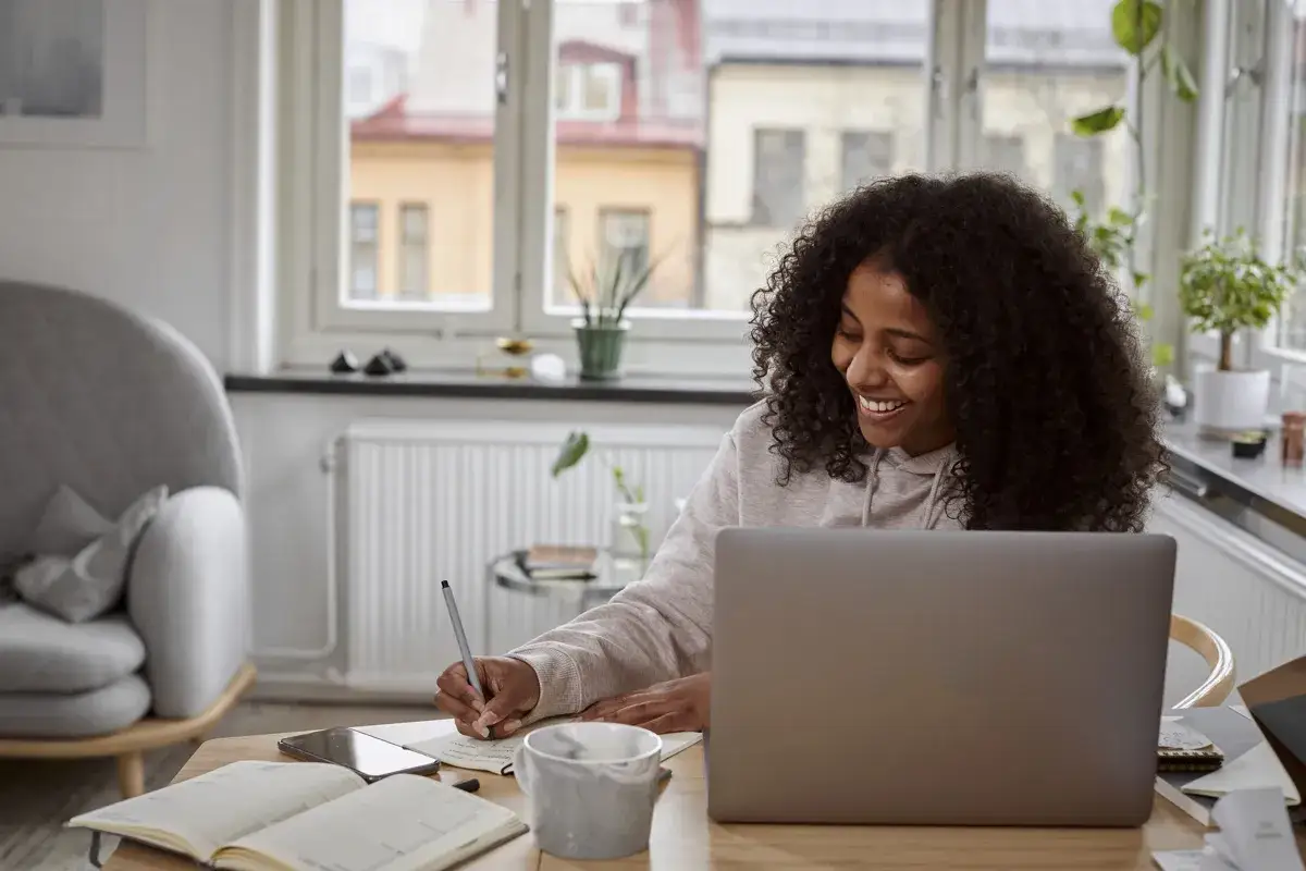 Glad kvinna med lockigt h&aring;r pluggar via e-learning utbildning vid sin laptop. Hon skriver anteckningar i en bok.