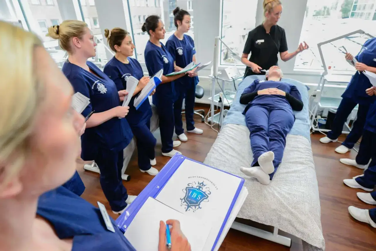 Elever p&aring; hudterapeut utbildning lyssnar p&aring; instruktioner. En elev ligger p&aring; en brits medan andra st&aring;r runt och antecknar.