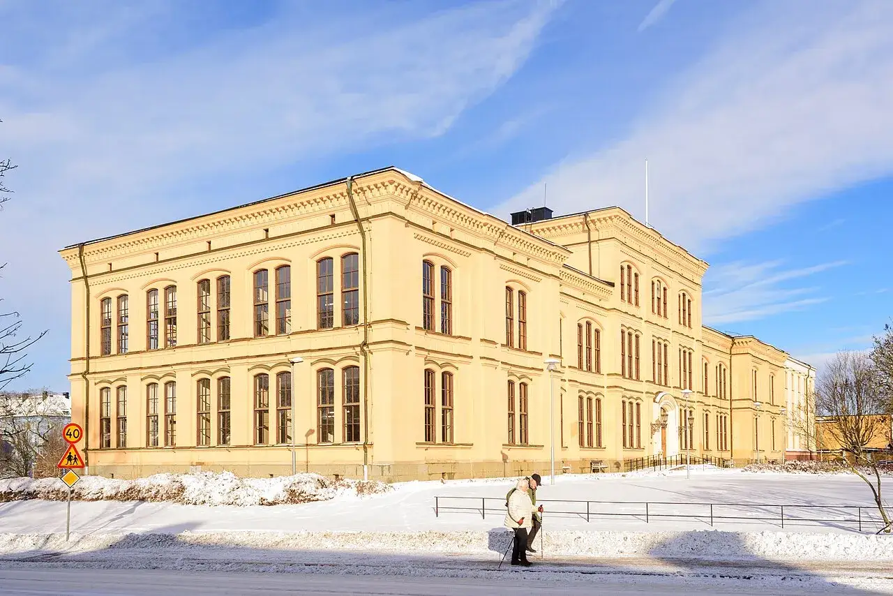 Katedralsskolan i Uppsala, en gul byggnad i vinterskrud med sn&ouml; p&aring; marken. Tv&aring; personer promenerar framf&ouml;r skolan.