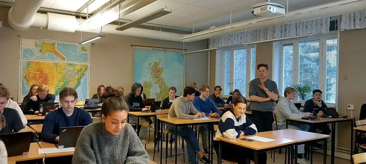 Elever p&aring; en arena academy sitter vid sina b&auml;nkar med laptops. En l&auml;rare st&aring;r och pratar framf&ouml;r klassrummet.