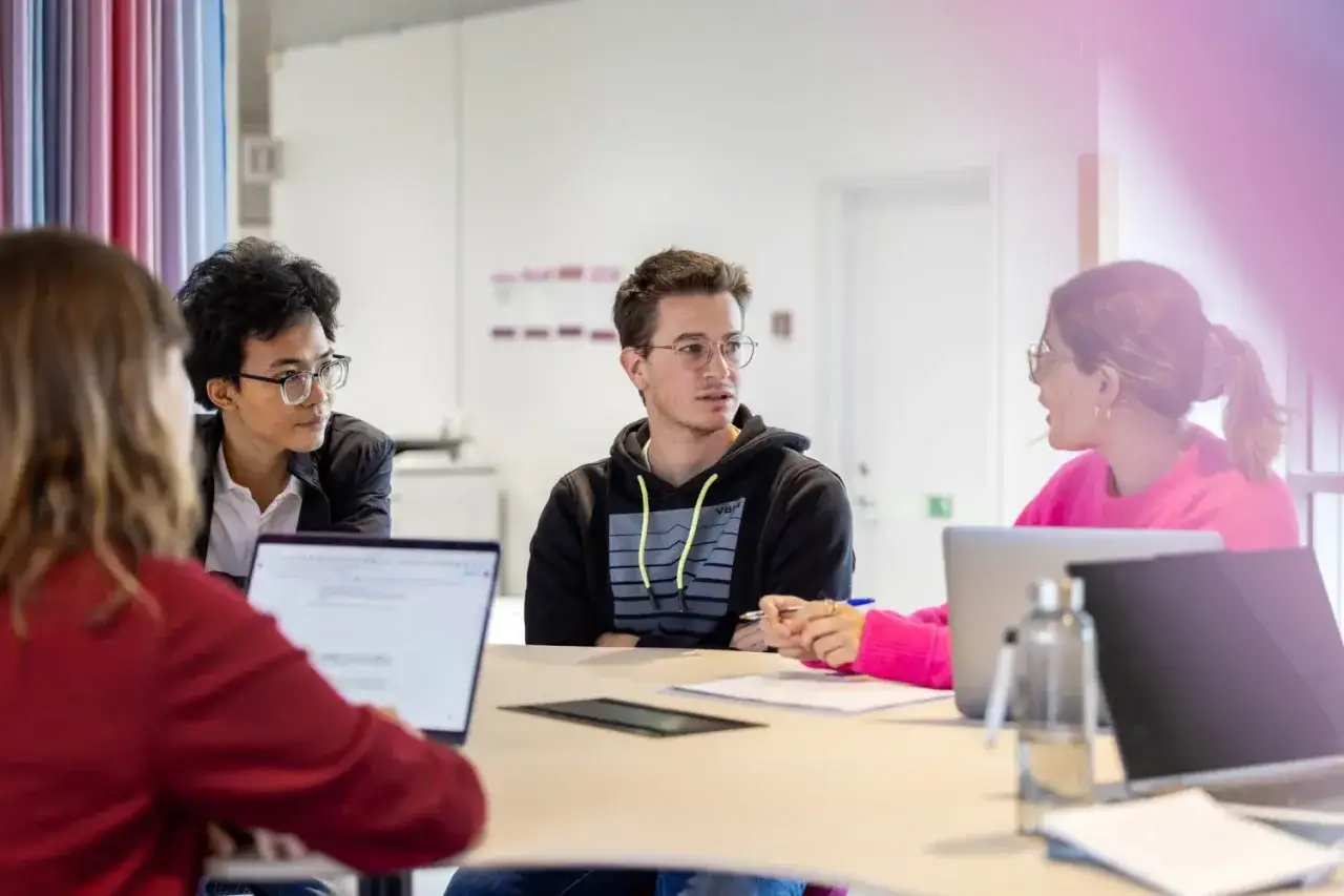 Fyra personer sitter runt ett bord med laptops. En kvinna i rosa pratar med en man i hoodie, en semistrukturerad intervju pågår.