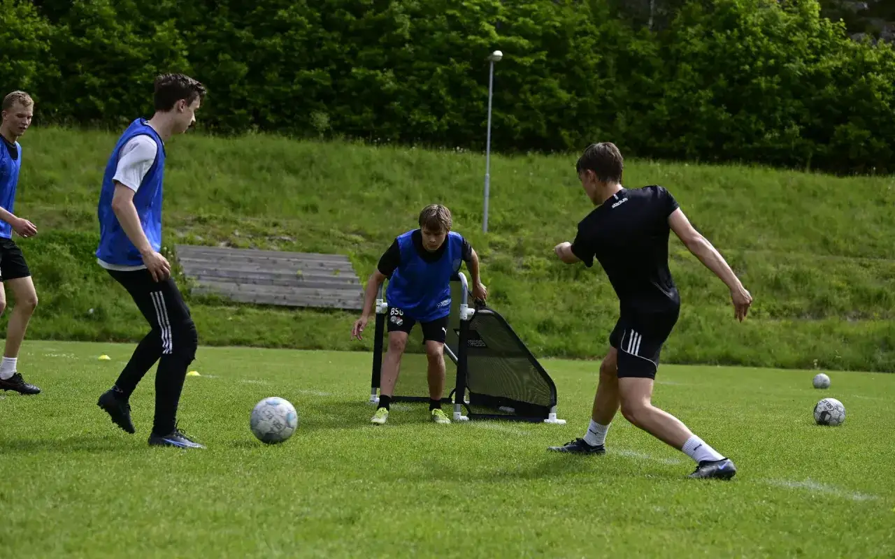 Unga spelare tr&auml;nar fotboll p&aring; en gr&ouml;n gr&auml;splan. En spelare i bl&aring; v&auml;st &ouml;var med en liten portabel m&aring;lbur.