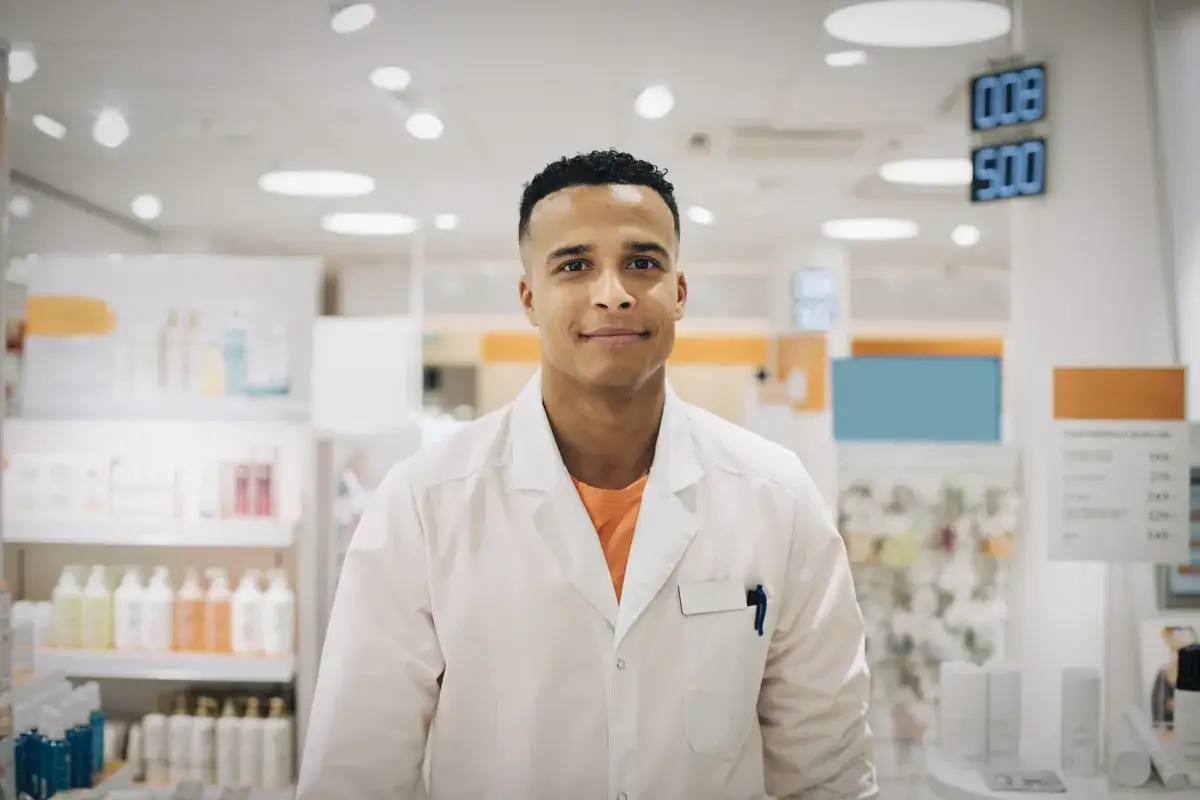 Ung man i vit rock, redo f&ouml;r apotekstekniker utbildning. Bakgrund med hyllor fyllda av produkter.