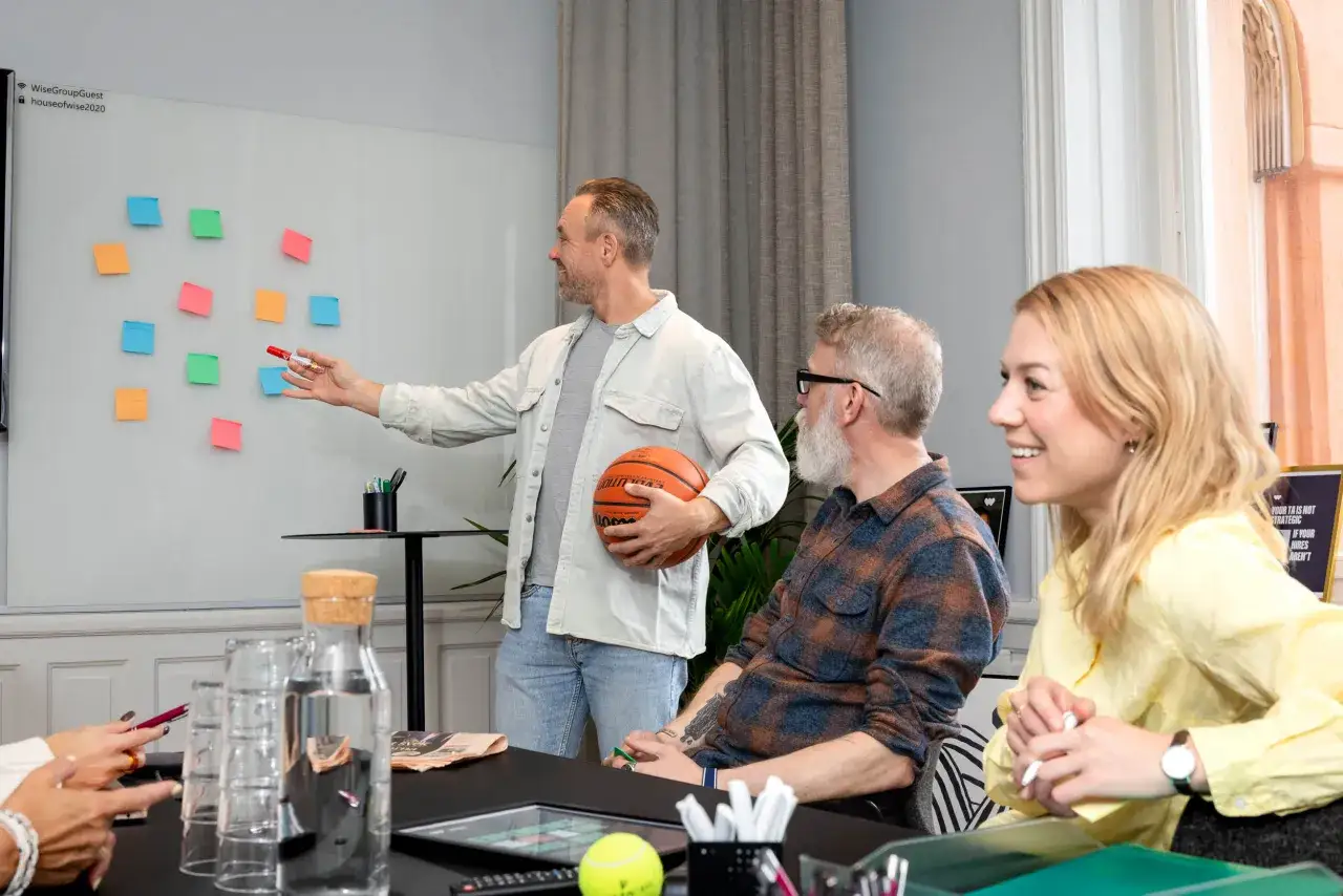 M&auml;n och kvinnor i m&ouml;tesrum diskuterar strategier med post-it-lappar och en basketboll. En digital strateg leder diskussionen.
