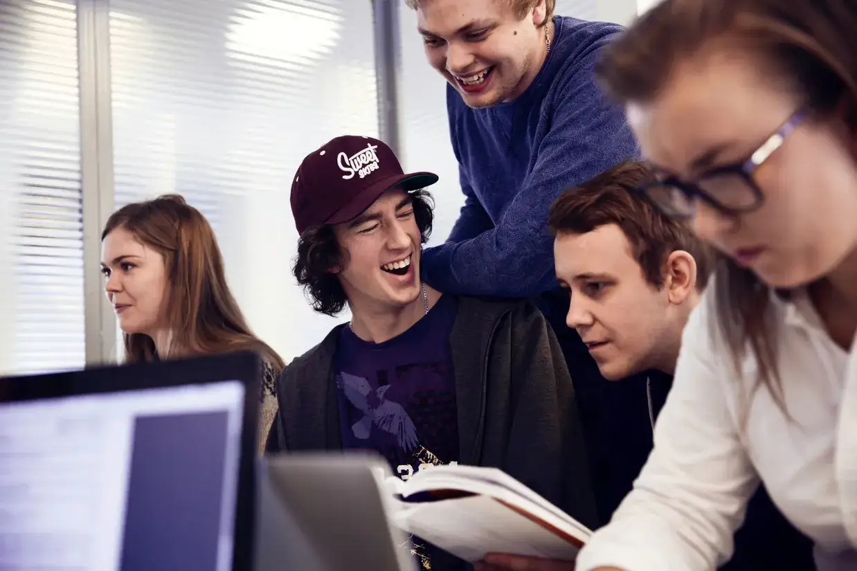 Studenter skrattar och samarbetar kring datorteknik 1a. En bok delas mellan tv&aring; killar, medan en tjej med glas&ouml;gon tittar ner.