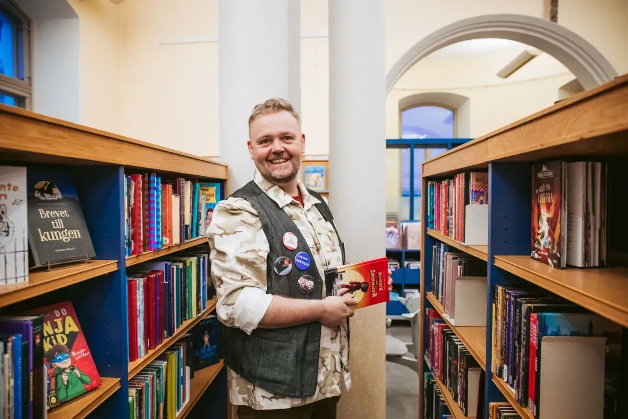 Glad man i bibliotek med böcker. Hans kläder och märken antyder en intressant bakgrund, kanske relaterad till bibliotekarie utbildning.