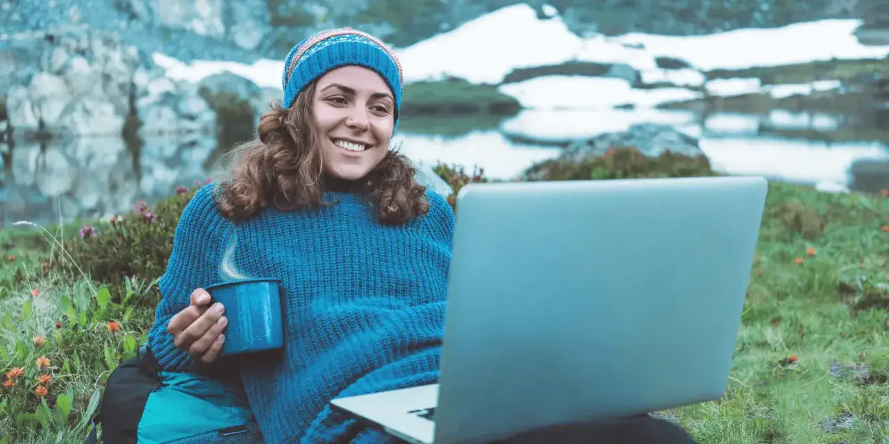 Kvinna med m&ouml;ssa och halsduk sitter utomhus med en laptop och en mugg varm dryck. Hon studerar engelska 6 distans vid en sj&ouml; med sn&ouml;kl&auml;dda berg i bakgrunden.