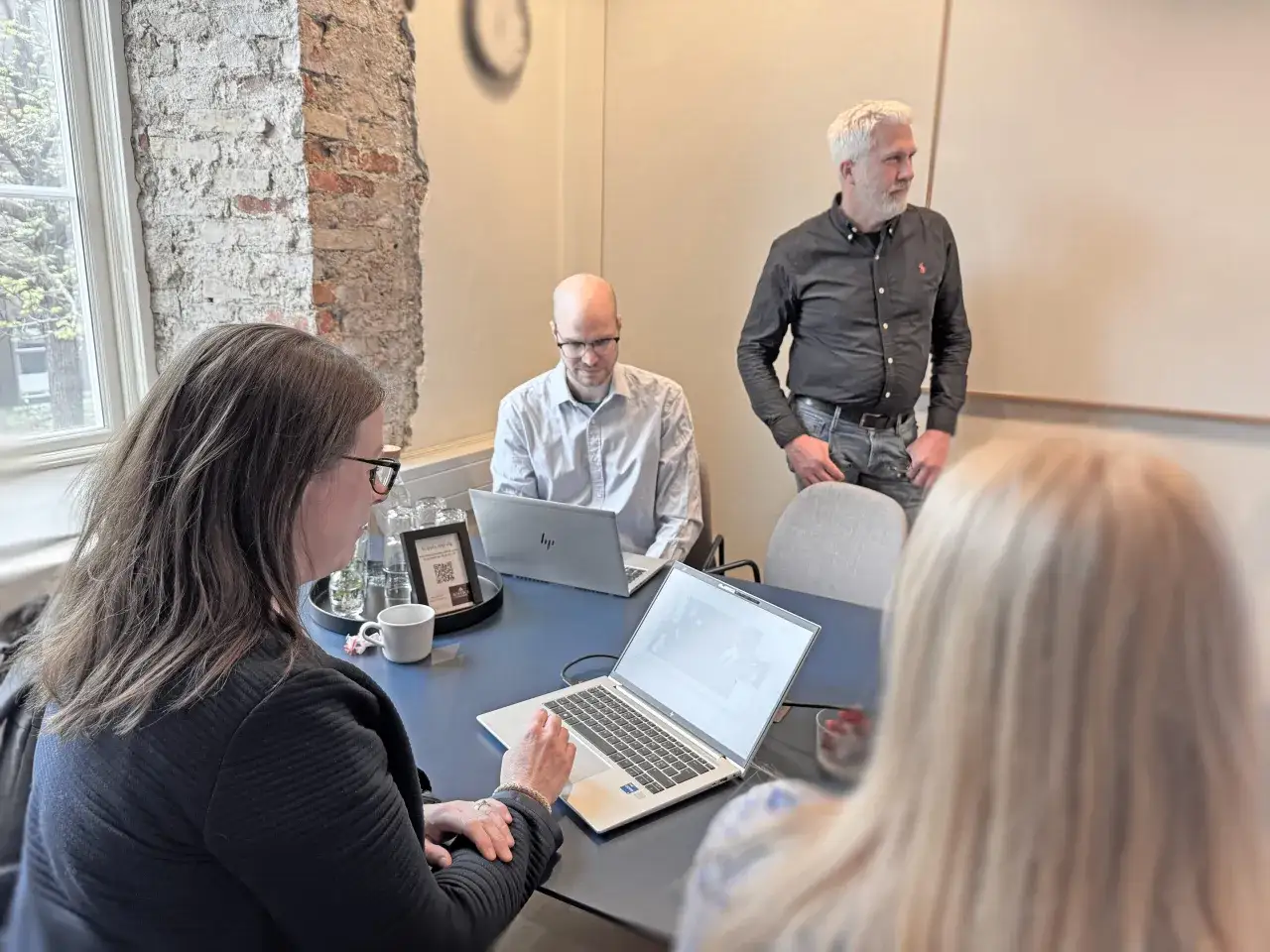 M&ouml;tesrum med tre personer. En kvinna med glas&ouml;gon arbetar vid en laptop. En man med glas&ouml;gon sitter vid en annan laptop. En man med gr&aring;tt h&aring;r st&aring;r och pratar. En teknisk s&auml;ljare diskuterar med teamet.