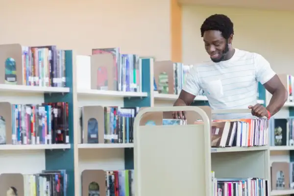 Man med bokvagn i bibliotek, kanske på väg att sortera böcker som en del av sin bibliotekarie utbildning.