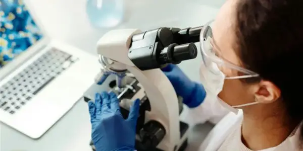 En forensiker arbetar med ett mikroskop i ett laboratorium. En laptop syns i bakgrunden.