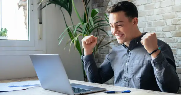 Ung man jublar framför sin laptop, kanske har han hittat roliga jobb eller lyckats med ett projekt.